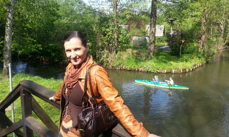 Kathrin Clara Jantke mitten im Spreewald. Sie steht auf einer Brücke, trägt eine braune Lederjacke und genießt den schönen Tag