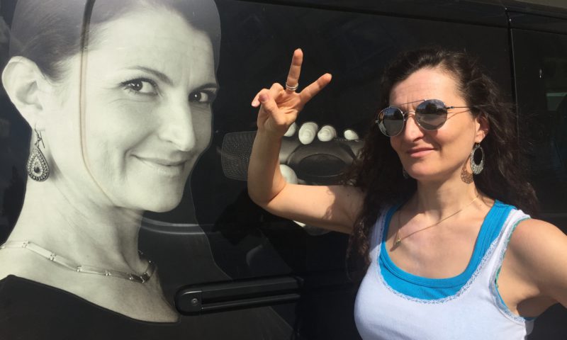 Kathrin Clara Jantke vor ihrem Auto auf dem ihr Gesicht in Überlebensgröße zu sehen ist. Sie schuat in die Kamera und macht ein Peace-Zeichen mit der rechten Hand. Sie trägt ein Sommeroutfit und eine Sonnenbrille